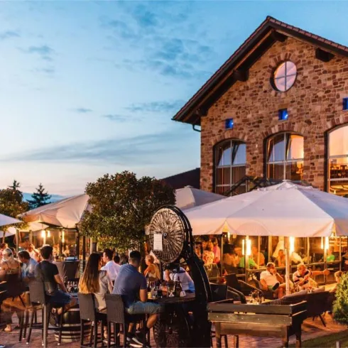 Eine beleuchtete Terasse mit Sonnenschirmen und Bestuhlung am Abend . Im Hintergrund ist ein großes,steiniges Gebäude zu sehen. 