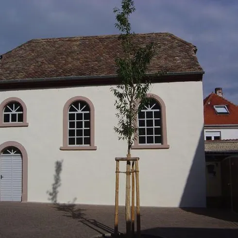 Die ehemalige Synagoge in Weisenheim am Berg. Die Synagoge ist ein weißes Gebäude mit runden Fenstern und Türen. das Foto ist aus seitlicher Sicht gemacht.Vor der Synagoge ist ein gepflasterter platz.