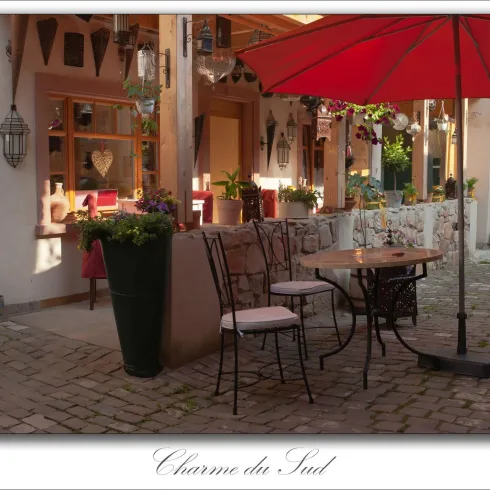 Im Vordergrund stehen Sitzmöbel und Tisch, zusammen mit einem Schirm. Dahinter ein einladendes Cafe und dessen Eingang.