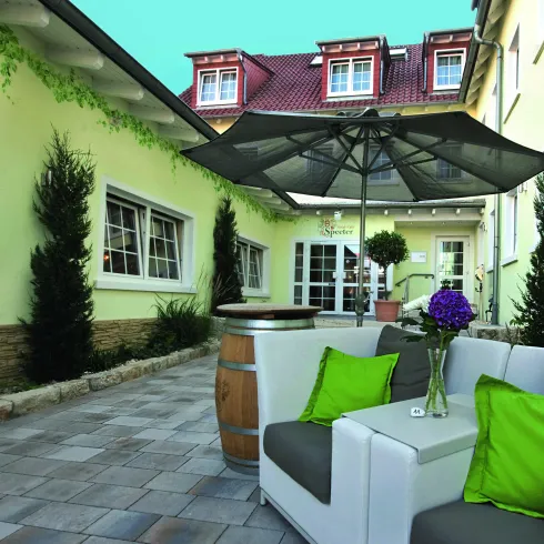 Ein gemütlicher Außenbereich mit modernen Möbeln und grünen Kissen. Im Hintergrund sind eine Terrasse und Pflanzen zu sehen.