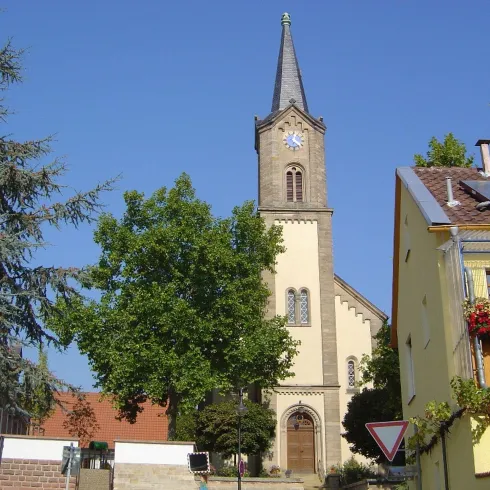 Prot. Kirche Erpolzheim, Eingangportal mit Kirchturm, Bäumen und Nachbarhaus. 