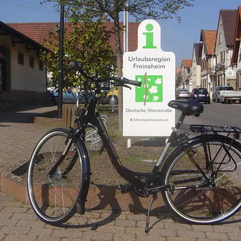 Ein schwarzes Fahrrad steht vor einem Informationsschild über die Urlaubsregion Freinsheim. Im Hintergrund sind Häuser und Bäume sichtbar.